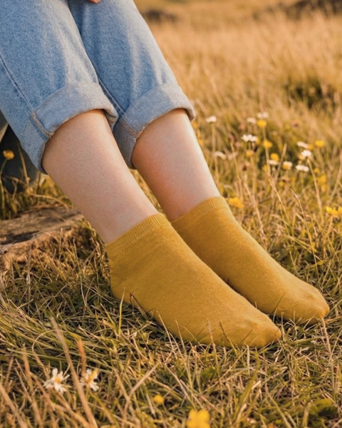 A close-up shot focuses on a person's lower legs resting in a grassy field dotted with small yellow and white wildflowers. They are wearing light blue cuffed denim jeans and mustard yellow ankle socks, bathed in warm golden hour light.