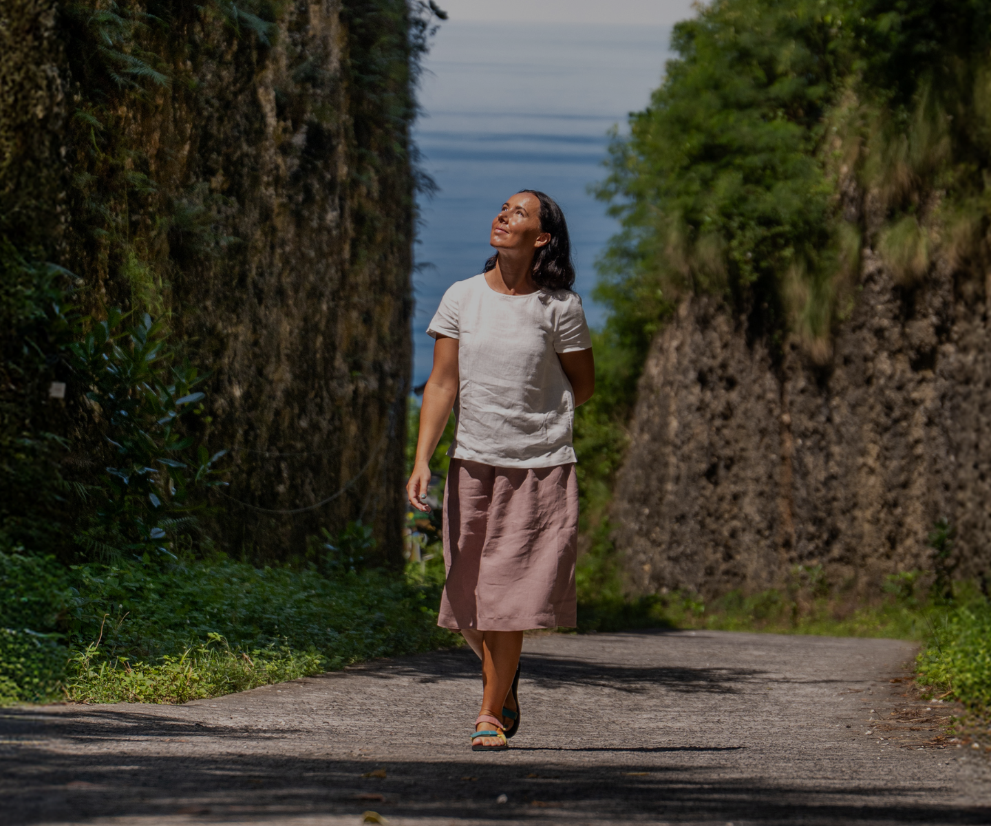A woman walks down a paved pathway nestled between two high, moss-covered stone walls, looking up towards the sky. She is dressed casually in a white short-sleeve top and a loose, dusty pink skirt.