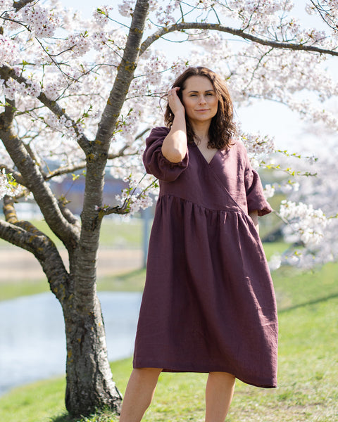 A person with medium-length dark hair stands in front of a cherry blossom tree, looking at the camera. They are wearing a long, shadow purple linen dress with a V-neck and short, loose sleeves. White cherry blossoms are visible on the branches of the tree behind them, and in the background, more blossoming trees line what appears to be a body of water or a path.