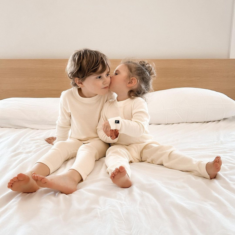 Two children wearing natural merino wool base layer sets, sitting on a bed.