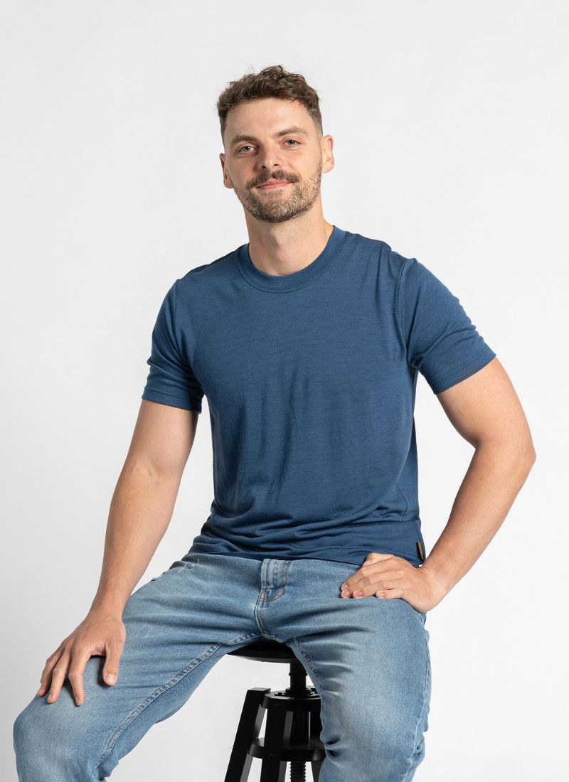 Man wearing a blue t-shirt and jeans sitting on a stool against a white background
