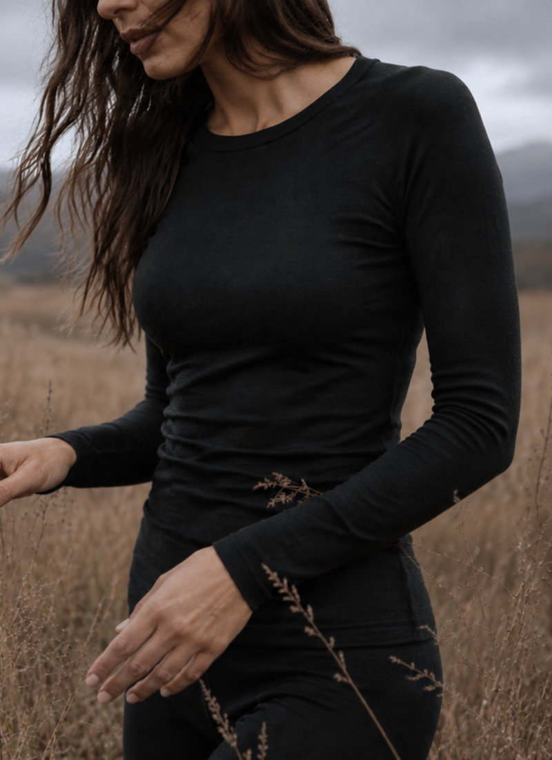 A woman with long wavy hair stands in a field of dry grass wearing a form-fitting black long-sleeved top and matching bottoms. She looks downward with a soft expression against a softly blurred backdrop of rolling hills and an overcast sky.