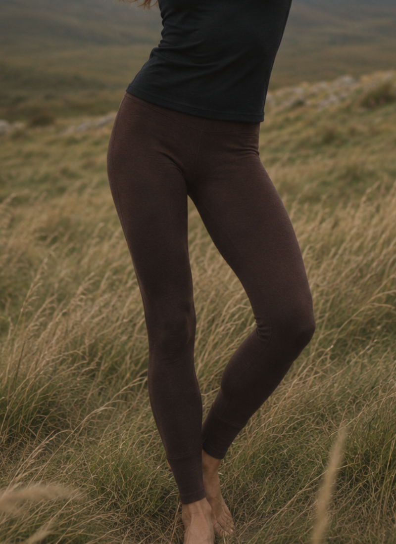A person stands barefoot in a field of tall, dry grass with their legs slightly crossed. The cropped image focuses on their dark brown form-fitting leggings and the lower hem of a black shirt.