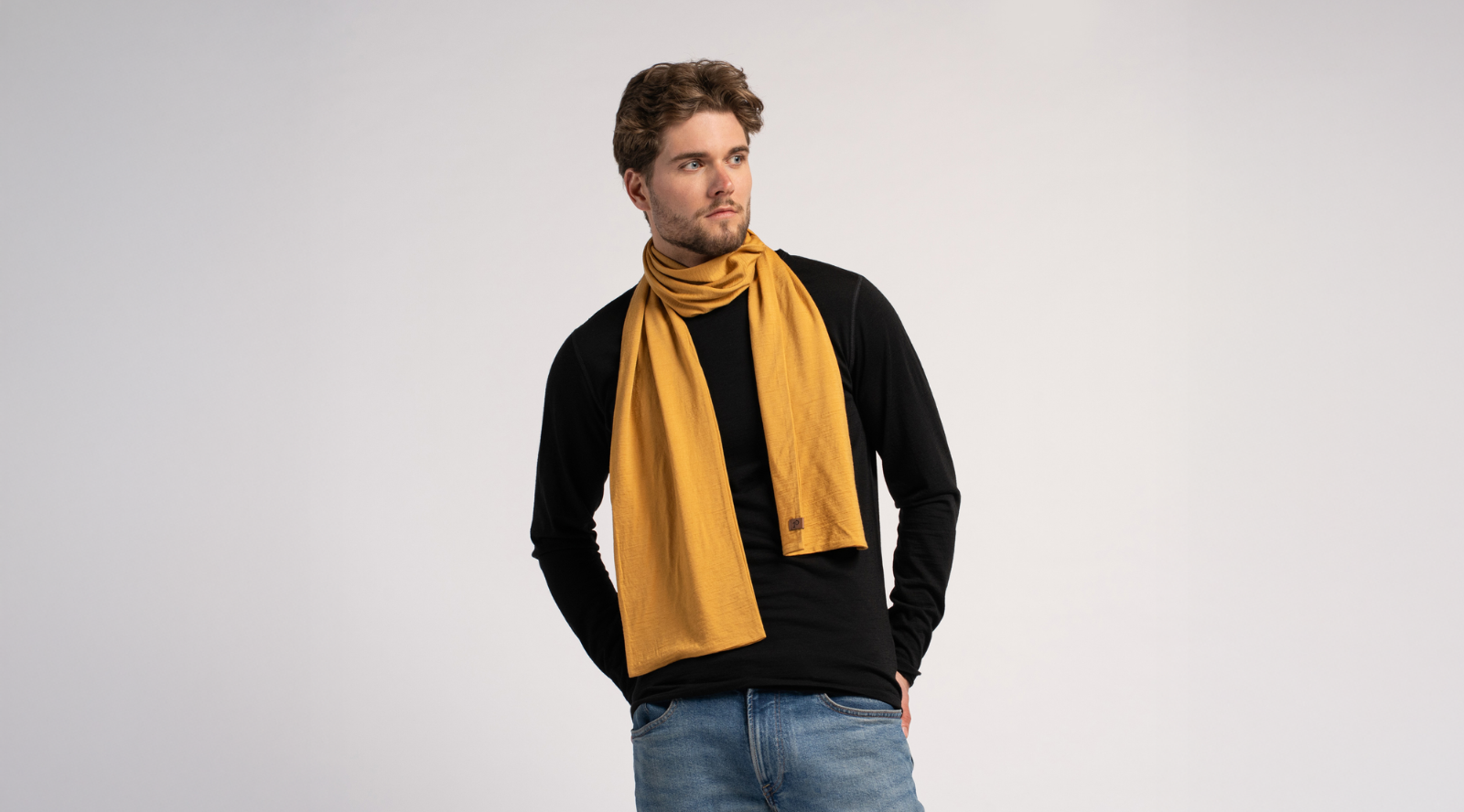 A man with a short beard stands against a light grey studio background wearing a mustard yellow knit scarf. He pairs the bright scarf with a black long-sleeved top and blue jeans while resting his hands in his pockets.