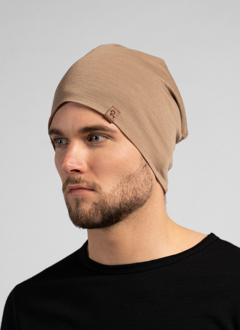 A bearded man looks slightly off-camera while wearing a slouchy, light brown knit beanie. He is dressed in a black crew-neck top against a seamless light grey studio background.