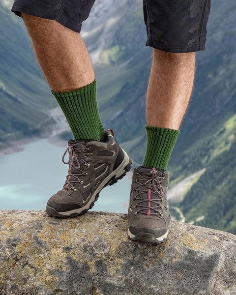 Front view of hiker wearing green ribbed Merino hiking socks with hiking boots, standing on a rock overlooking mountains and lake.