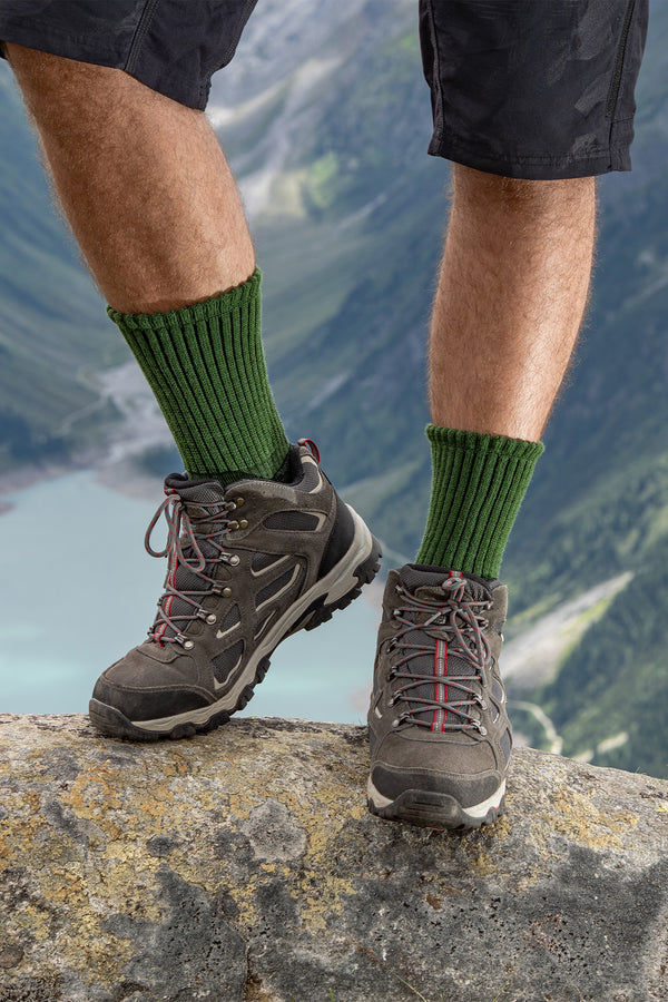 Front view of hiker wearing green ribbed Merino hiking socks with hiking boots, standing on a rock overlooking mountains and lake.