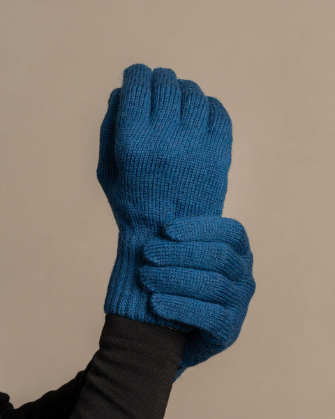 
A close-up shot of a person&#39;s hands wearing menique brand vibrant blue, ribbed knit gloves. The left hand is clenched into a fist, while the right hand appears to be gripping the wrist area of the left glove. The person is wearing a dark long-sleeved shirt underneath. The background is a solid, neutral gray.