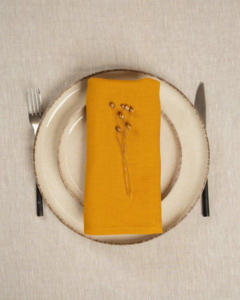 The menique linen napkin in spicy yellow color, put on top of two stacked plates . There are cutlery items by the sides of the plates. The background is a natural color linen tablecloth.