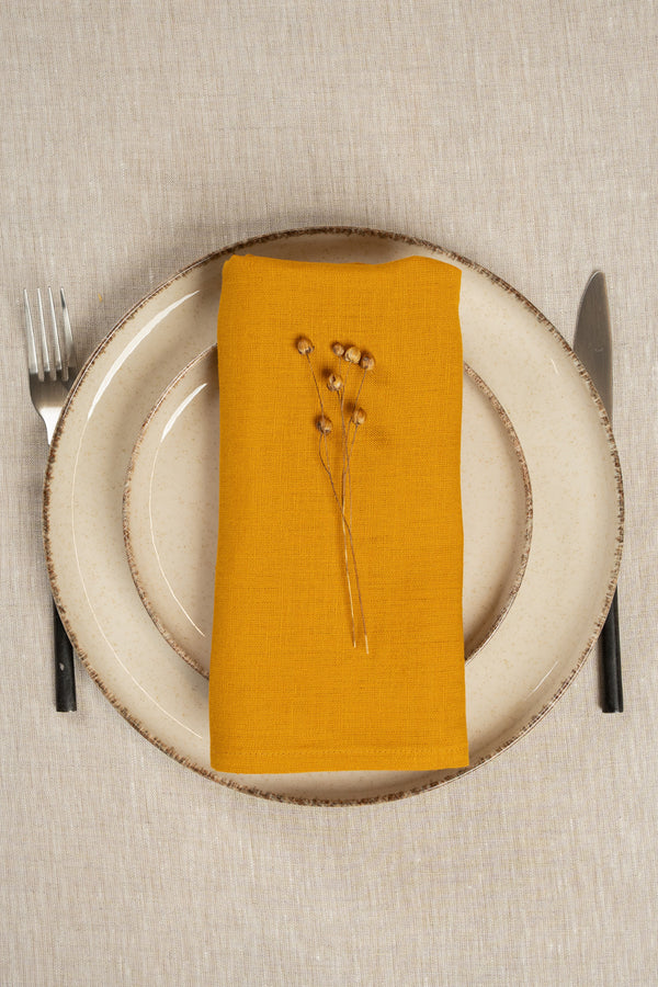 The menique linen napkin in spicy yellow color, put on top of two stacked plates . There are cutlery items by the sides of the plates. The background is a natural color linen tablecloth.