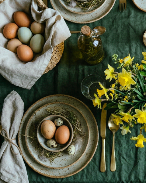 An elegant flat-lay view of a dark forest green linen tablecloth. The sophisticated arrangement includes speckled stoneware plates holding a small "nest" of eggs, a light linen napkin tied with rustic twine, vintage-style gold cutlery, and a vibrant bunch of yellow daffodils.