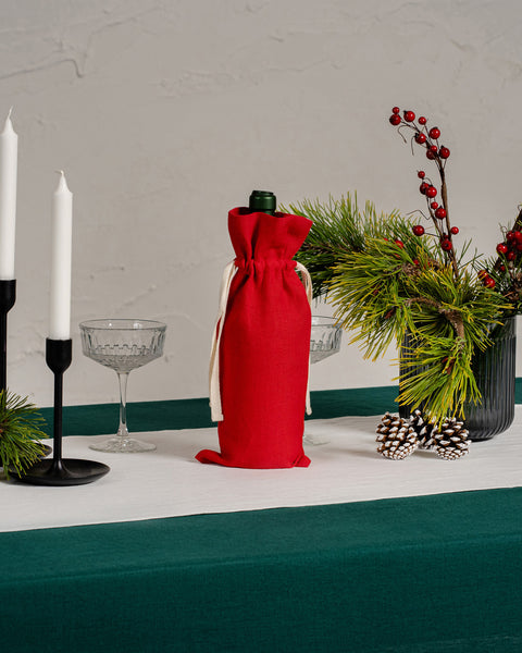 Elegant Christmas table décor featuring a dark green linen tablecloth, white linen table runner, red linen wine bag, glassware, black candle holders, pine greenery, red berries, and pinecones.