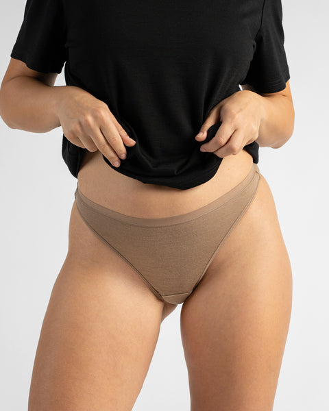Front view of a woman lifting the edge of a black cropped t-shirt, showcasing beige low-rise cheeky underwear against a light backdrop.