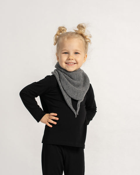 Smiling child wearing a dark grey knit triangle scarf over a black outfit, standing confidently against a light background.