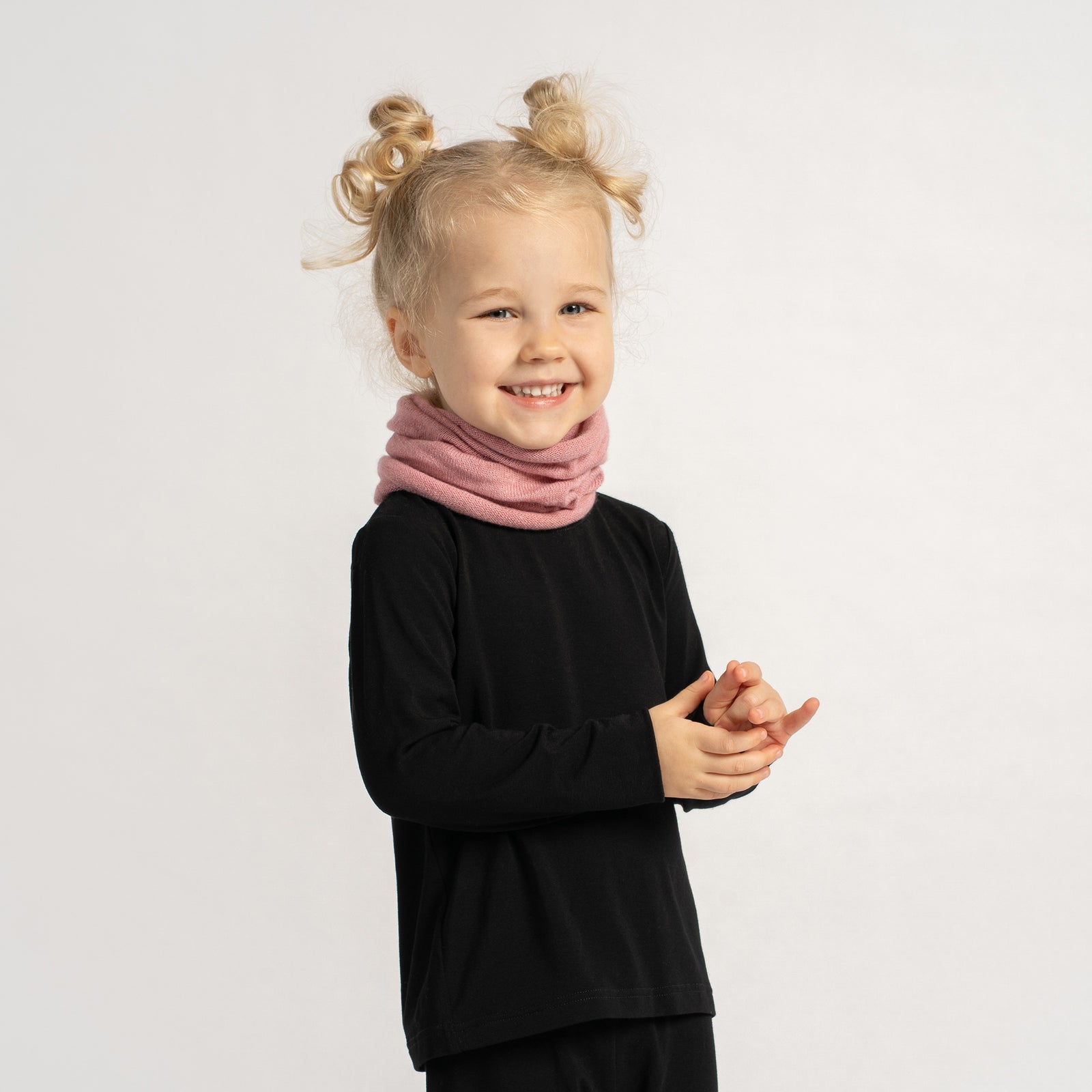 Little girl in a black outfit wearing a soft pink knit neck warmer, smiling with arms crossed.
