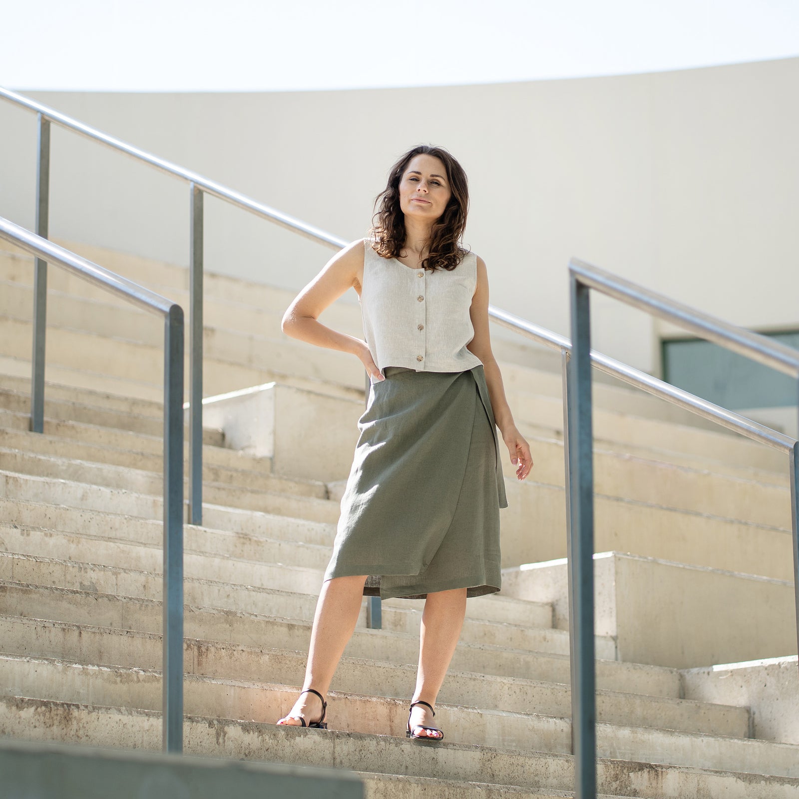 A person with short, curly dark hair stands in profile, looking down at their waist. They are wearing a white sleeveless top and a knee-length black wrap skirt with a long tie hanging down the front. On their feet are black sandals with multiple straps and a low block heel. To their left are two large potted plants against a light-colored wall.