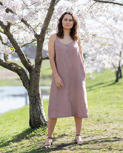 A woman with long, dark, wavy hair, wearing a sleeveless, button-front, faded rose midi-dress and black open-toed sandals, stands outdoors in front of a tree with white blossoms. She has her hands casually at her sides. She looks directly at the viewer with a neutral expression or slight smile. The background is a sunny, outdoor setting with green grass and a body of water, possibly a pond or lake, and more blossoming trees.