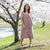 A woman with long, dark, wavy hair, wearing a sleeveless, button-front, faded rose midi-dress, stands outdoors in front of a tree with white blossoms. She holds a sprig of the blossoms up to her nose with her right hand. Her left hand is on her hip. She looks directly at the viewer with a slight smile. The background is a sunny, outdoor setting with green grass and more blossoming trees.