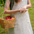 A woman with long red hair wearing a natural color linen apron, standing in a lush green garden and holding a woven basket.