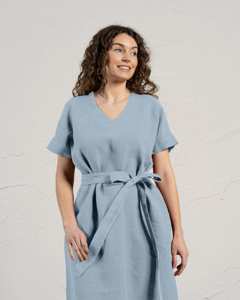 A woman with curly brown hair smiles while modeling a cloudy blue linen menique dress with short sleeves and pockets.