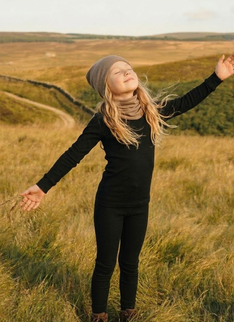 A blonde child standing in a sunlit, grassy field with arms joyfully outstretched, wearing a black long-sleeved top and dark pants.