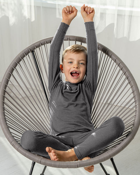 Smiling boy in gray Merino wool base layer set sitting cross-legged on a chair with arms raised.