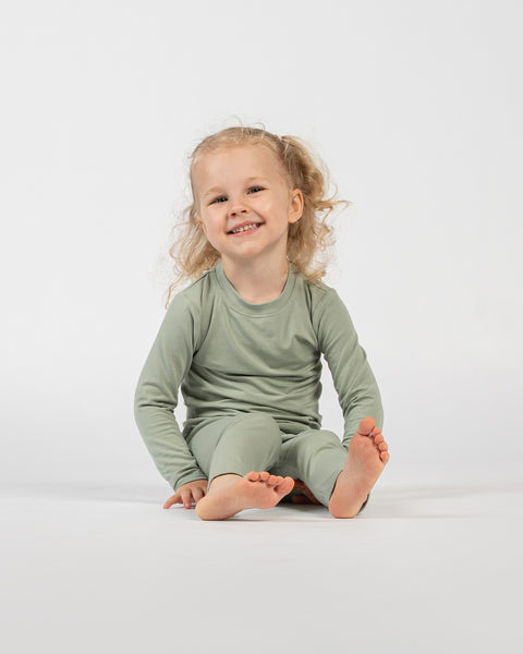 Child sitting barefoot in green bamboo long-sleeve top and bottoms, smiling.