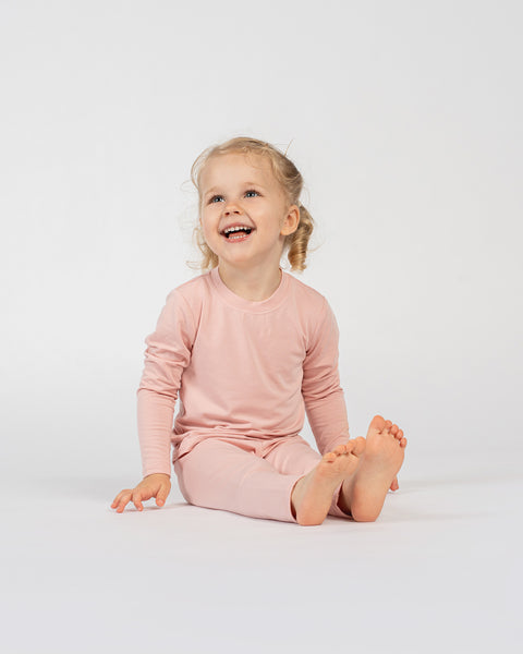 Smiling child sitting on the floor in pink bamboo long-sleeve top and bottoms.