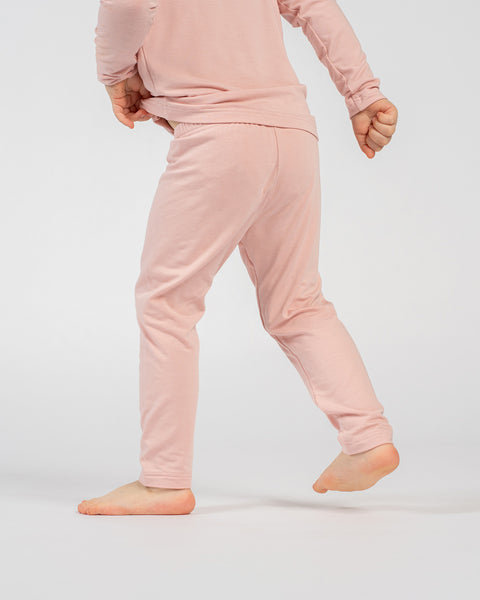 Child in pink long-sleeve top and matching pants, standing barefoot with back facing the camera and arms slightly bent, showing the outfit from behind against a plain background.