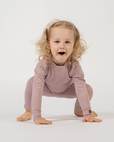 A child with blonde curly hair and a light pink floral headband is in a crouching position on a white background, wearing a dusty pink long-sleeved top and matching pants. The child has their hands on the floor and is looking at the camera with an open-mouthed expression.