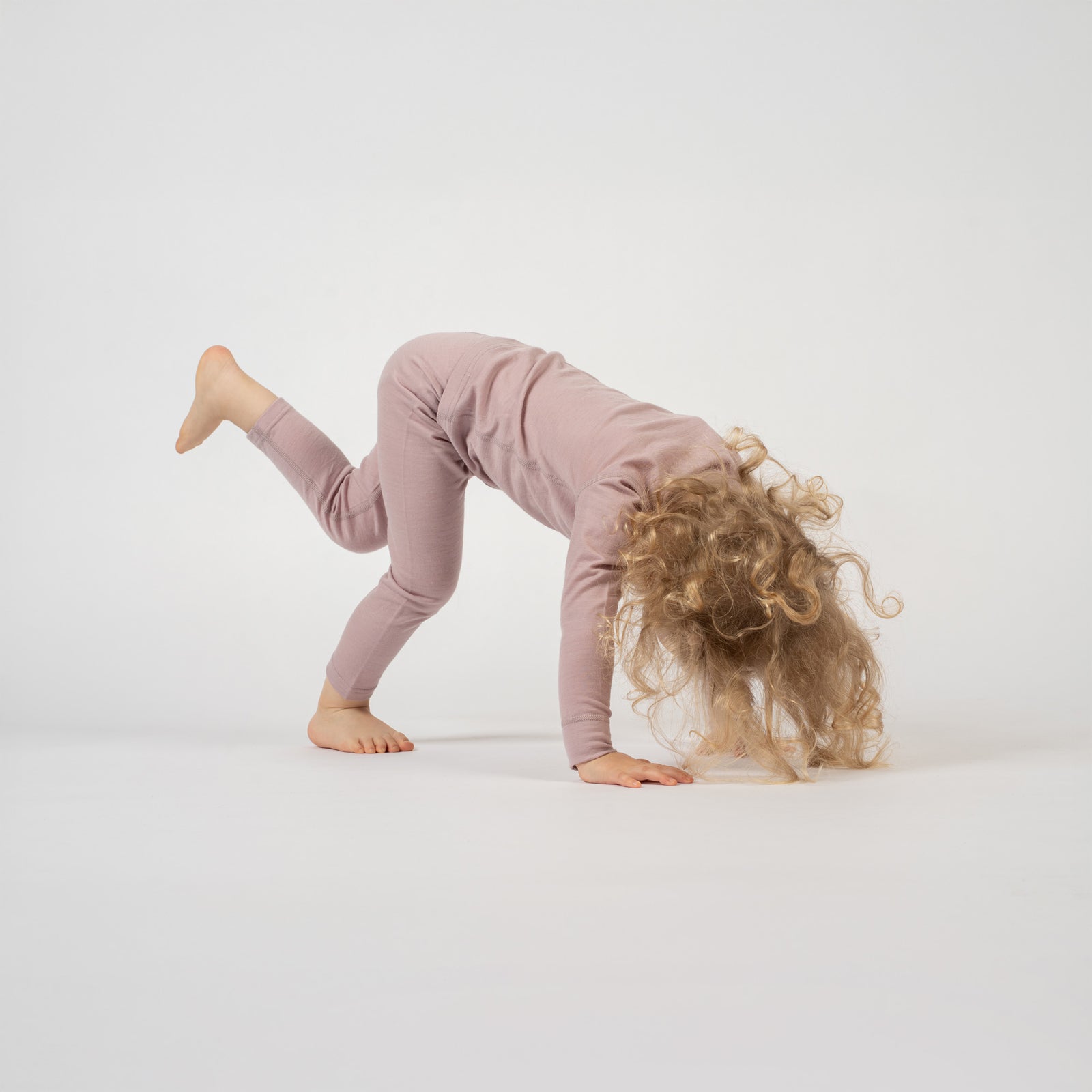 A smiling child with curly blonde hair and a light pink floral headband sits cross-legged on a white background, wearing a dusty pink long-sleeved top and matching pants. The child is looking directly at the camera.