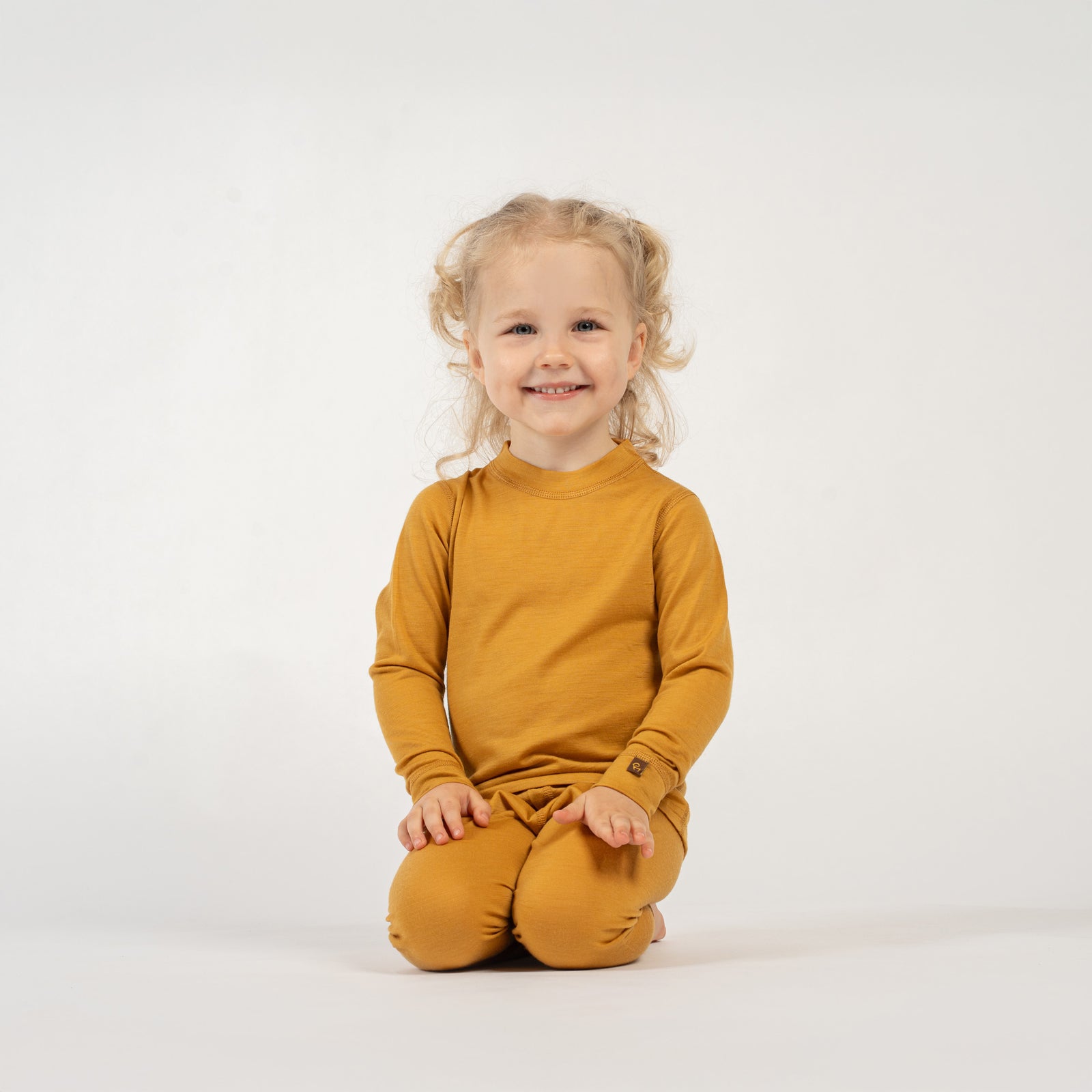 A playful child with curly blonde pigtails, wearing a yellow long-sleeved top and matching pants, is bending over with their hands on their hips, looking up at the camera with a wide smile. The child is barefoot on a white surface.
