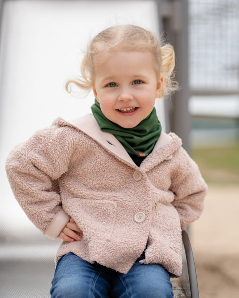 A young child with blonde hair and blue eyes smiles brightly while sitting on a slide, wearing a beige Menique jacket and a green neck warmer. The child&#39;s cheerful expression suggests a fun moment outdoors.