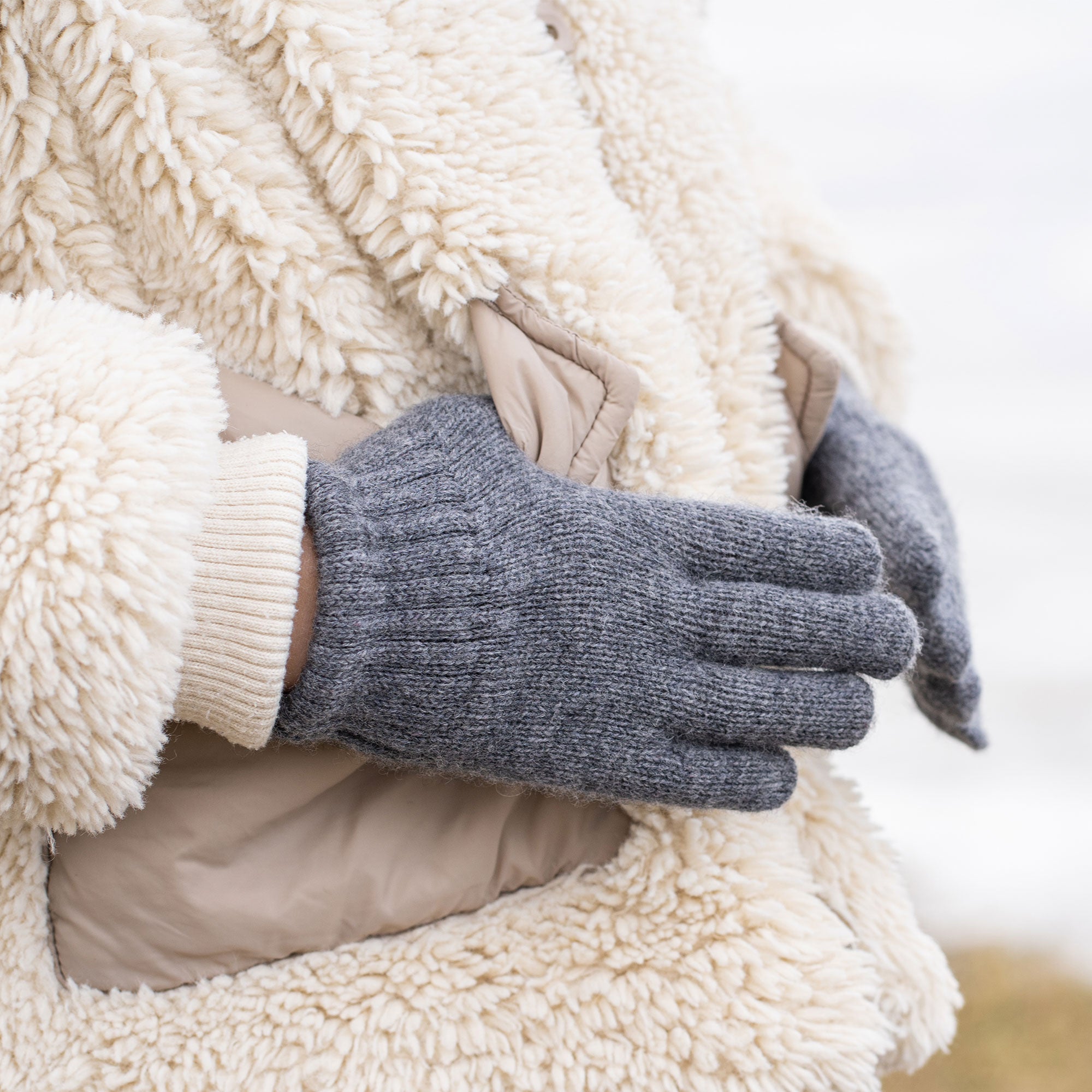 A person wearing a cream-colored sherpa coat with beige pockets and a light-colored ribbed cuff is putting on or adjusting a dark grey knitted glove. The background is a blurred, light outdoor scene.