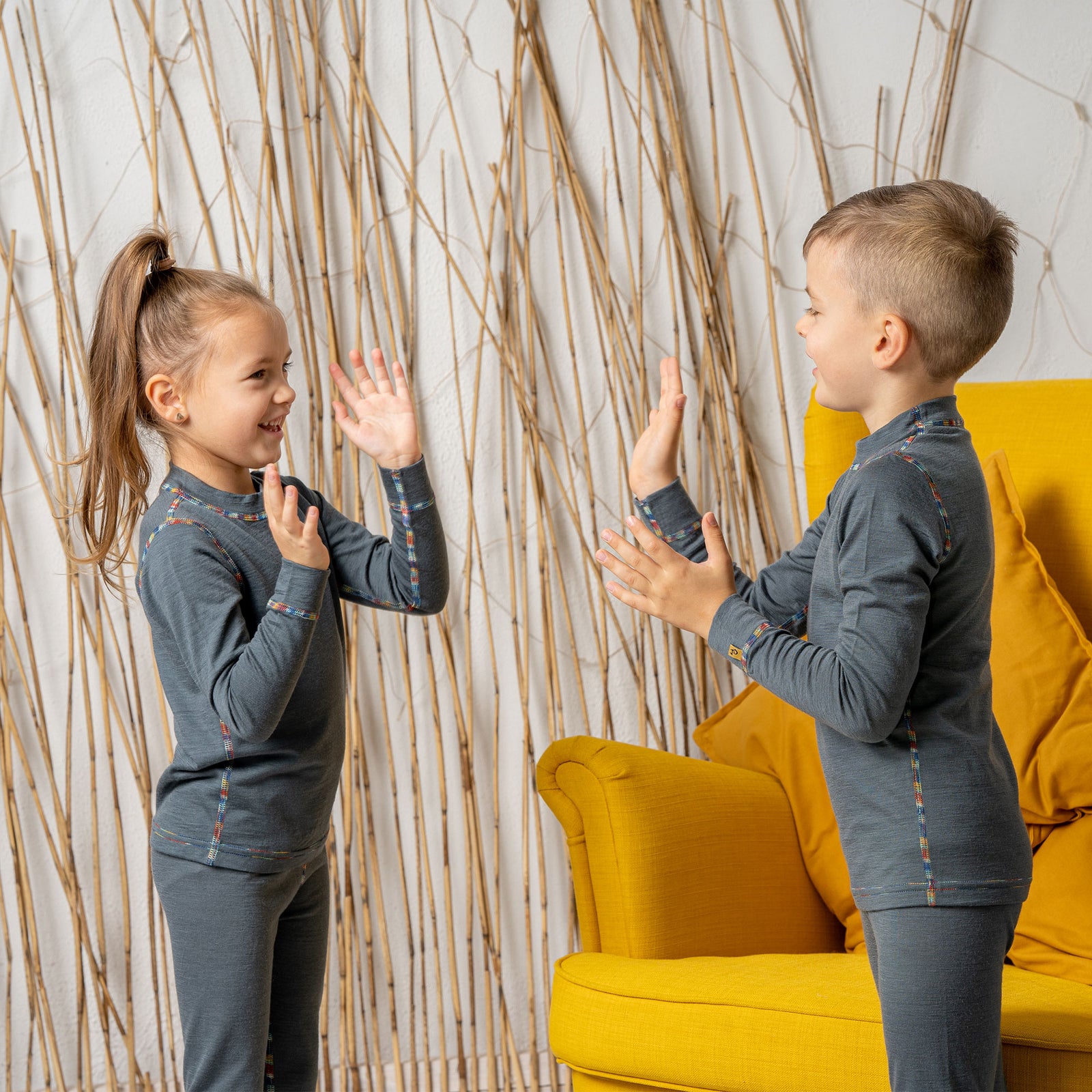 A boy sitting in a yellow armchair, wearing the menique Kids' 160 Merino Pants in Perfect Grey color. He has his hands raised above his head, being cheerful.