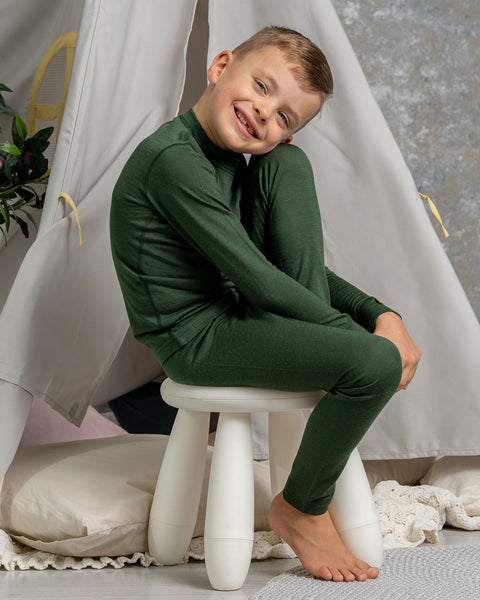 A boy sitting an a chair, with one leg on top of it, wearing the menique Kids&#39; 160 Merino Pants in Dark Green color. He is resting his head on the raised leg&#39;s knee.