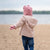 A joyful child wearing a pink balaclava with ears, a pink scarf, and a beige coat stands on a beach, hands open in excitement.