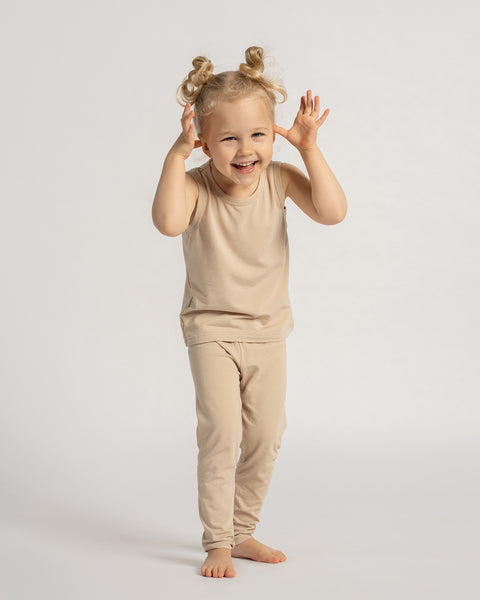 Smiling toddler wearing a beige sleeveless top and leggings set, standing barefoot and playing in front of a light background.