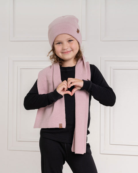 A child with long, wavy hair wears a dusty pink menique knit beanie with a small brown tag, a pink knit scarf, and a black long-sleeved shirt and black pants. They are standing indoors in front of a white wall with decorative molding. The child is looking at the camera with a smile, and their hands are brought together in front of their chest to form a heart shape.