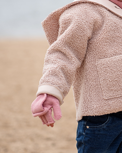 A child wearing a pink sherpa coat, pink gloves, and blue jeans stands on a sandy beach.