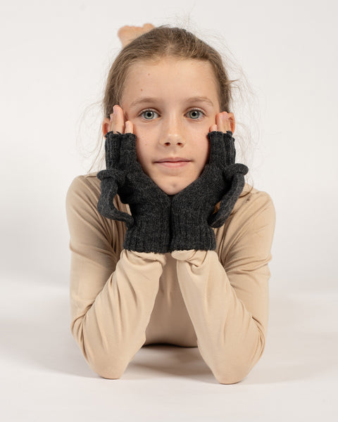 Girl lying down with head resting on hands, wearing dark gray knitted convertible mittens with mitten flaps open.