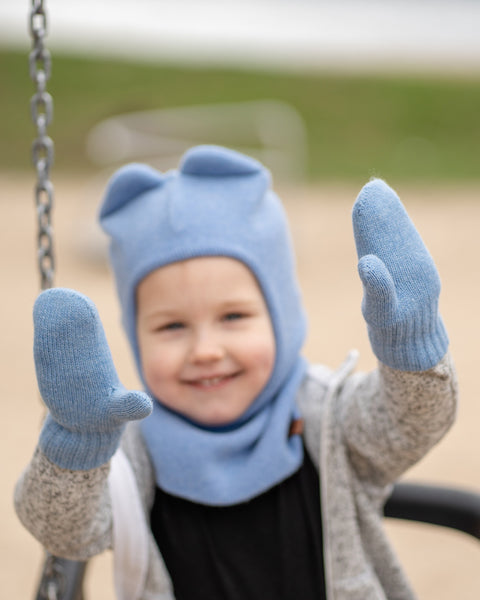 A young child wearing a light blue balaclava with small ears, light blue mittens, and a grey and black outfit, smiles while looking at the viewer. They are partially obscured by a metal swing chain on the left, with a blurred sandy ground in the background.