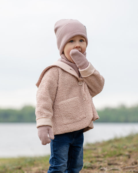 A young child is standing outdoors with a hand near their mouth. They are dressed in a light pink knit beanie with a small brown tag, a light pink or beige textured jacket with large buttons and a collar, and blue jeans. They are also wearing matching dusty pink menique mittens. In the background, there is a body of water under a light, possibly overcast, sky.