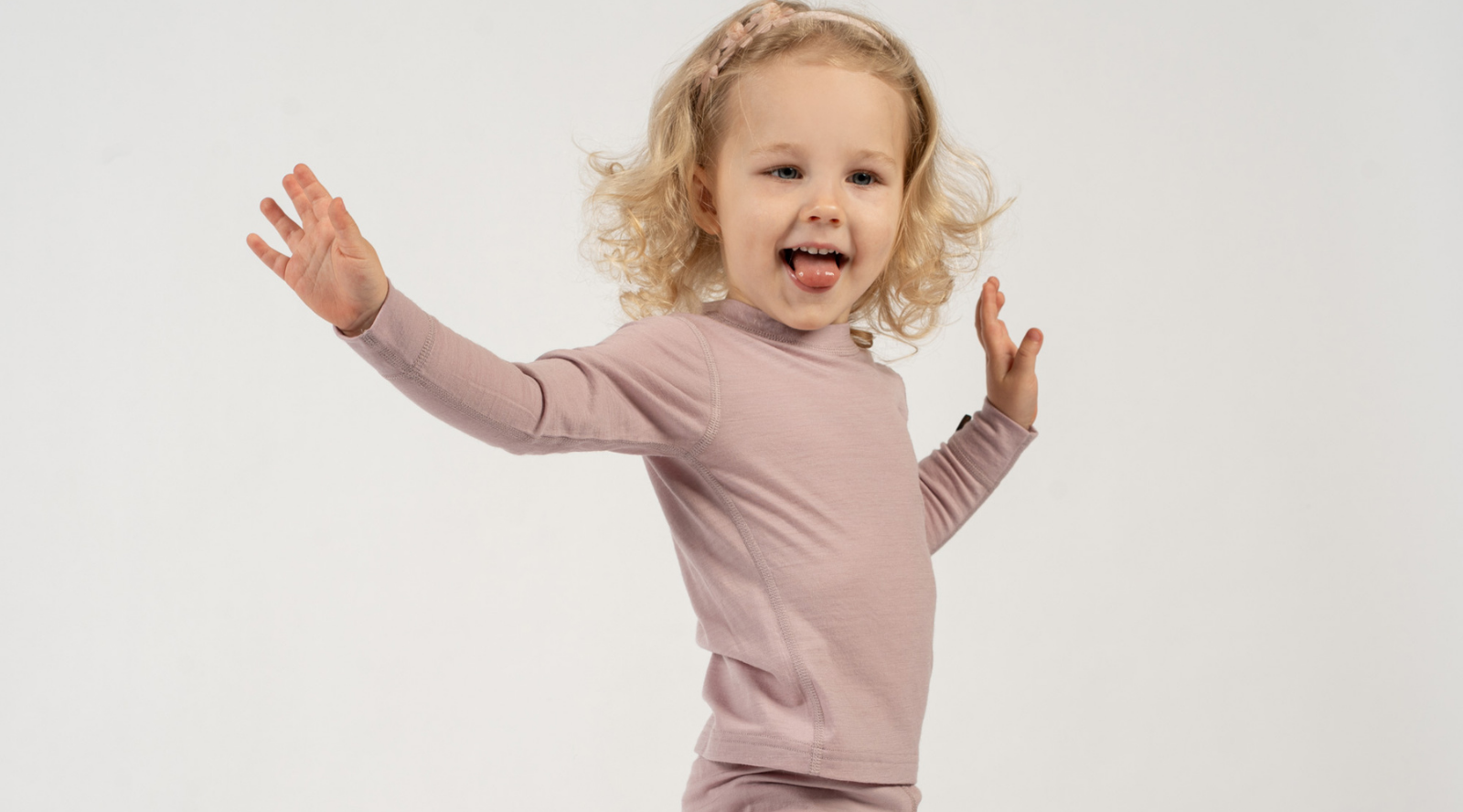A young girl with curly blonde hair and a small headband stands playfully against a plain white background. She has her arms thrown wide open with an excited expression and her tongue slightly out, wearing a dusty mauve long-sleeve top and matching bottoms.