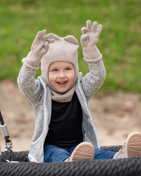 A cheerful young child, with blue eyes and an open-mouthed smile, sits on a black rope swing with arms raised. They are wearing a menique creamy beige balaclava with ears, a matching scarf, beige gloves, a grey speckled zip-up hoodie, blue jeans, and pink sneakers. The background is blurred green grass and brown dirt.