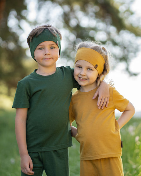 Two children standing outdoors wearing matching Merino wool T-shirts, shorts and headbands in yellow and dark green. The lightweight, breathable kids' outfits are showcased in a sunny park setting, perfect for everyday play, family outings, and warm-weather adventures.
