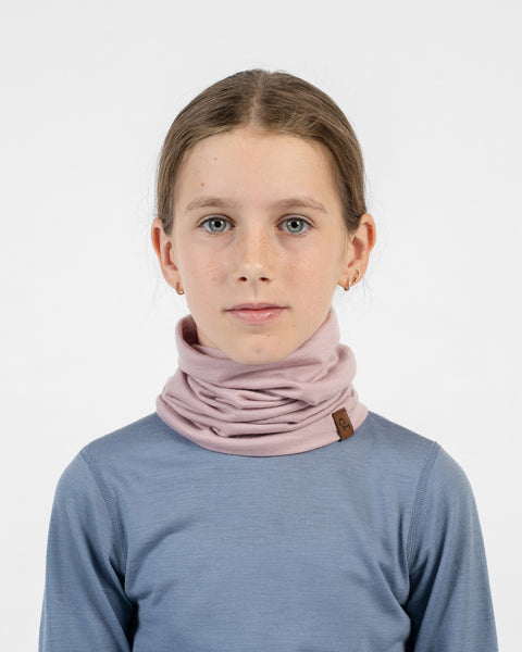 A close-up front view of a young girl wearing a dusty pink merino long neck gaiter bunched around her neck below her chin, paired with a dusty blue long-sleeve shirt.