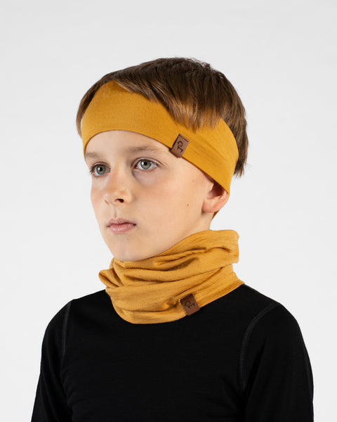 A close-up studio portrait of a young boy looking off to his right against a plain white background. He is wearing a black long-sleeved top, a mustard yellow headband, and a matching mustard yellow neck gaiter bunched loosely around his neck.