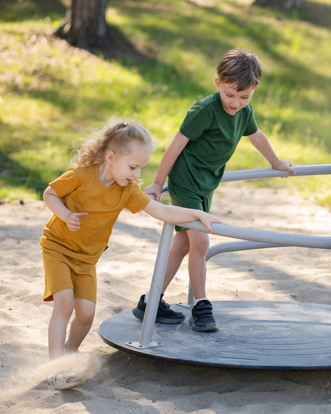 Two children  outdoors wearing matching Merino wool T-shirts and shorts in yellow and dark green. The lightweight, breathable kids' outfits are showcased in a sunny park setting, perfect for everyday play, family outings, and warm-weather adventures.