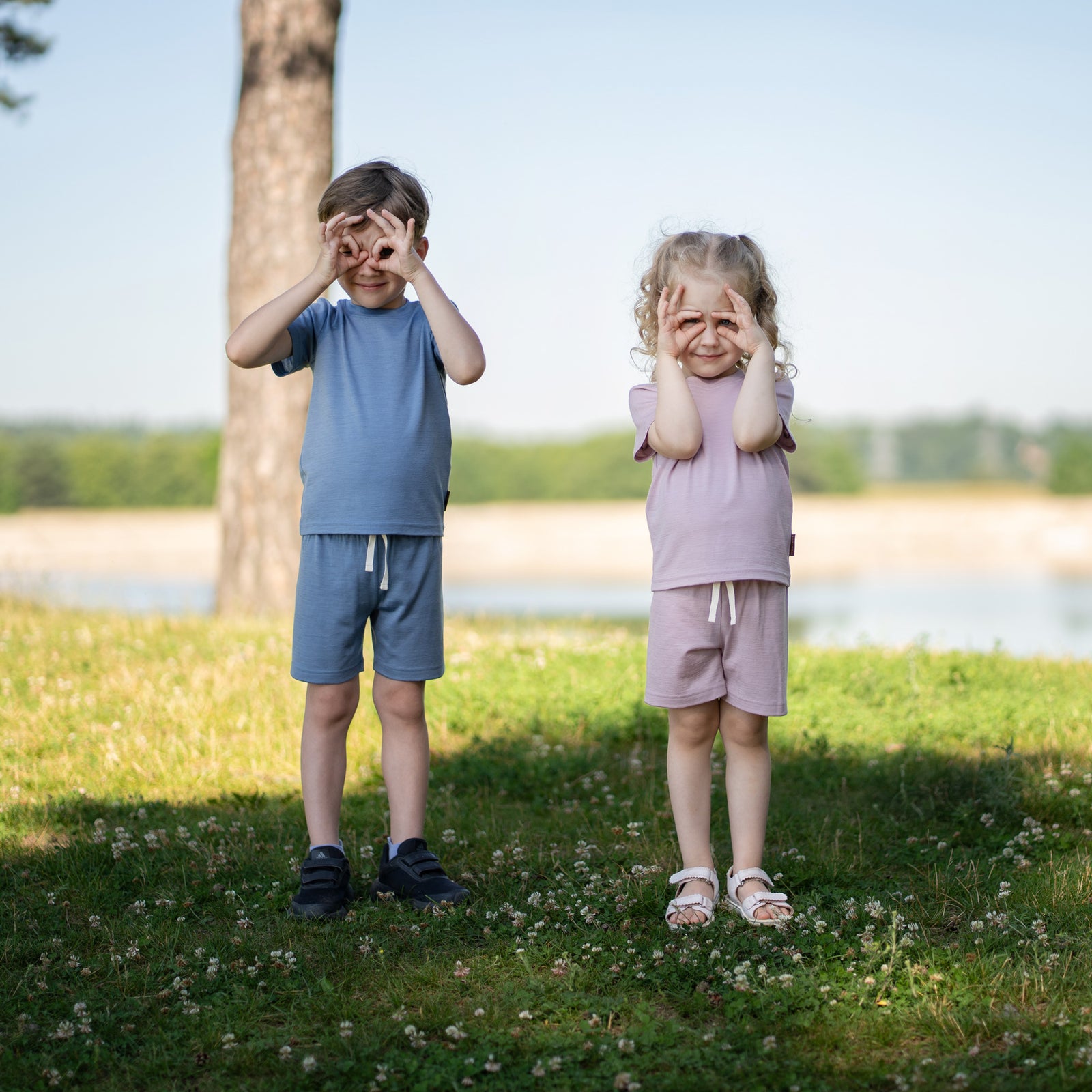 Young boy sitting cross-legged on a wooden park bench wearing a dusty blue Merino wool T-shirt and matching shorts. The soft, breathable kids' outfit is showcased in a sunny outdoor setting, ideal for everyday comfort and warm-weather adventures.